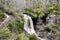 A scenic waterfall roadside going over the cliff down to the boulders and rocks below where you can no longer drive under but can