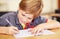 School, education and a student boy writing in a book while sitting at his desk in a class room for learning. Kids