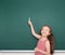 Schoolgirl child in red striped dress point and drawing on green chalkboard background, summer school vacation concept