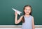 Schoolgirl with paper plane play near a blackboard, empty space, education concept