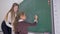 Schoolgirl with piece of chalk writes an example on the blackboard with help of a female teacher at the math lesson