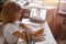 Schoolgirl writes listening to teacher at videocall via modern laptop at table in light room