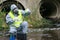 A scientist in a gray protective suit and mask, on a pond, holding test tubes and a flask of blue and green liquid