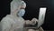 Scientist man in a protective suit and mask typing on a laptop night writes patient reports in laboratory