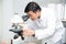 Scientist using Microscope in Laboratory. Male Researcher wearing white Coat sitting at Desk and looking at Samples.