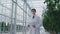 A scientist in a white coat with a table in his hand in the middle of the greenhouse with tomatoes