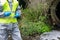 Scientist in yellow protective vest, uniform and gloves, collects plant materials from the river with tweezers in a Petri dish,