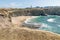 Sea waves reach the sand and cliffs of Ponta do Trovao coastline with buildings under the blue sky