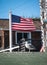 Seagull on the top of a green shingle roof with a blurred American flag flying in the background.