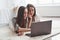 Searching for some good movie. Young female twins lying on the floor near white bed and using silver colored laptop