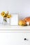 Seasonal autumn fall decorations with photo frame, colorful calendula flowers in a vase and pumpkins on the white chest of drawers