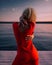 Seductive woman in a luxurious red dress stands near the river. Atmospheric sunset on the pier.