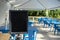 Selective focus on an empty black sign in front of a reserved seating area filled with blue chairs under an outdoor canopy