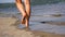 Selective focus footage of a human on a beach walking on the sand past the ocean waves.