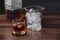 Selective focus on a glass with whiskey and three ice cubes. Defocused ice bucket, cap and ice bucket. Wooden table