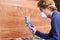 Selective focus on hands of young Asian worker with safety mask painting a piece of wood with spray gun in home workshop. Shallow