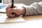 Selective focus at human hand using pen to write down short note on paper on working table. With blurred laptop computer at the