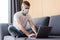 Selective focus of teleworker in medical mask using laptop while sitting on couch