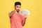 Self confident rich man with beard in striped t-shirt holding fan of dollars and pointing finger at camera with serious expression