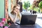 Self employed freelancer Caucasian woman working with her phone and laptop in a restaurant terrace in the street in a sunny day