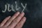 The semi-erased word JULY on the black chalkboard. An adult man`s left hand removes the handwritten word with his fingers. Fuzzy