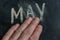 The semi-erased word MAY appear on the black chalkboard. An adult man`s left hand removes the handwritten word with his fingers.