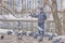 Senior Asian man in a jacket, jeans and a knitted hat feeds birds in a city park near the river in winter. Doves are sitting on