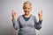 Senior beautiful grey-haired woman wearing golden queen crown over white background shouting with crazy expression doing rock