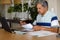 Senior biracial man doing paperwork and using laptop in dining room