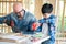 A senior carpenter and boy wearing goggle in the modern wood workplace. A man looking at boy who used screwdriver. Young child