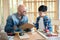 Senior carpenter and kid wearing goggle in the modern wood workplace Man holding clipboard and talk with a boy. Boy point at paper