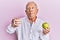 Senior caucasian man holding denture and green apple making fish face with mouth and squinting eyes, crazy and comical