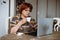 Senior concentrated woman sitting at table on white chair, holding cup of coffee, looking at laptop, working from home.