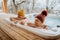 Senior couple in kintted cap enjoying together outdoor bathtub at their terrace during cold winter day.