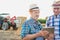 Senior farmer standing while using digital tablet with mature farmer against farm tractor, round bale hay in field with y