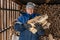 Senior farmer in a warm hat and jacket holds firewood against the background of a woodpile in the open air
