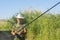 Senior fisherman in hat with a fishing rod prepares to fish on the shore with reeds. An elderly retired man is resting in nature.