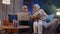 Senior grandparents couple reading book, using laptop pc on couch in night living room at home