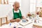 Senior grey-haired artist man smiling happy painting pottery sitting on the table at art studio