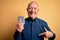 Senior grey haired man holding pharmaceutical pill drugs over yellow background with surprise face pointing finger to himself