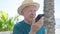 Senior grey-haired man tourist wearing summer hat sending voice message by smartphone at seaside