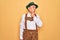 Senior grey-haired man wearing german traditional octoberfest suit over yellow background with hand on chin thinking about