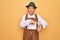 Senior grey-haired man wearing german traditional octoberfest suit over yellow background In hurry pointing to watch time,