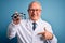 Senior grey haired optic doctor man holding optometrist eyeglasses over blue background with surprise face pointing finger to