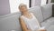 Senior grey-haired woman finds comfort in relaxation on cozy sofa, holding paper indoors in her sunlit living room, showcasing a