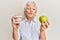 Senior grey-haired woman holding green apple and denture teeth looking at the camera blowing a kiss being lovely and sexy