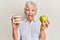 Senior grey-haired woman holding green apple and denture teeth sticking tongue out happy with funny expression