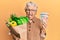 Senior grey-haired woman holding groceries and canadian dollars sticking tongue out happy with funny expression