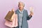 Senior grey-haired woman holding shopping bags and uk pounds banknotes in shock face, looking skeptical and sarcastic, surprised