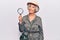 Senior grey-haired woman wearing explorer hat holding magnifying glass looking positive and happy standing and smiling with a